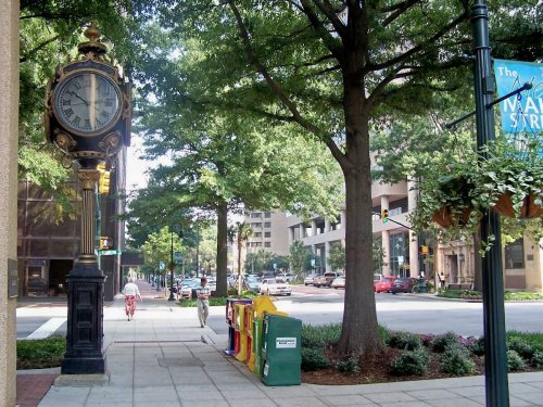 A street view featuring a vintage clock, trees, and newspaper stands along a sidewalk in a city.