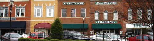 A row of brick buildings featuring shops, including a pharmacy and a drug store, with parked cars in front.