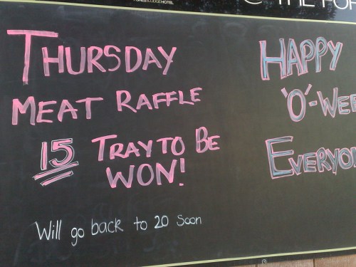 Chalkboard sign announcing a Thursday meat raffle with details about winning trays and a festive greeting.