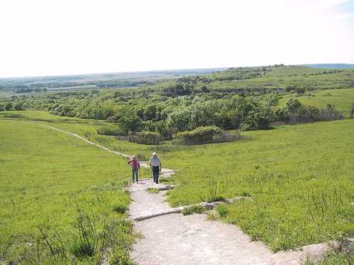 Two people walk along a rocky path through a lush green landscape under a bright sky.