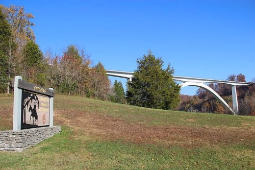 A scenic view of a bridge above a grassy area with a park sign in the foreground and trees in the background.