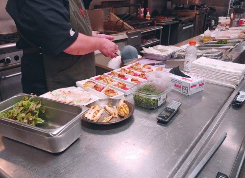 A chef prepares small dishes in a busy kitchen, with various ingredients and utensils on a stainless steel countertop.