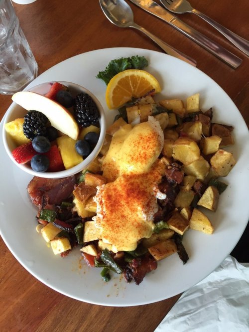 A plate of breakfast featuring eggs Benedict on potatoes, with a side of fresh fruit salad.