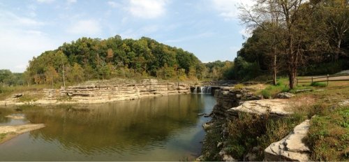 A serene landscape featuring a calm river, rocky cliffs, and lush green trees under a clear blue sky.