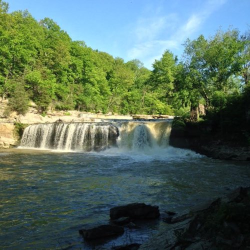 A serene waterfall cascades over rocks, surrounded by lush green trees and a clear blue sky.