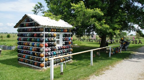 A colorful, whimsical cabin made of stacked bowling balls, set in a grassy area with trees and a clear sky.