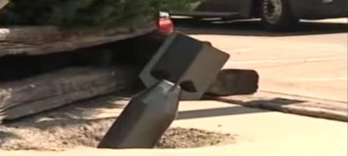 A large, black metal object is partially embedded in the ground near a wooden structure and a parking area.