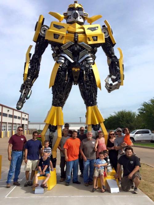 A large yellow robot sculpture stands with a group of people, including children, posing in front of it.