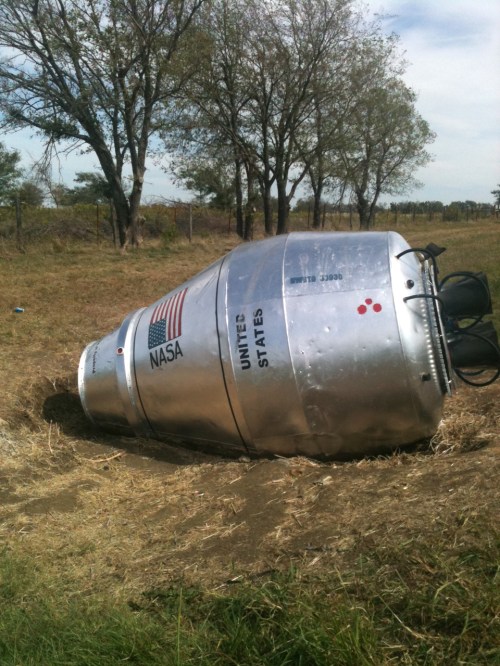 A silver NASA capsule lies on its side in a grassy field, surrounded by trees and open land.