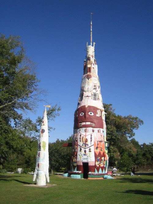 Colorful, tall totem-like structure with intricate designs, surrounded by greenery under a clear blue sky.
