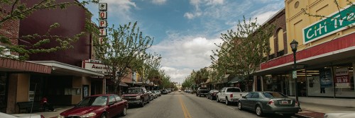 A quiet downtown street lined with trees, shops, and parked cars under a partly cloudy sky.