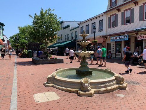 A decorative fountain in a bustling street with shops and people enjoying a sunny day.