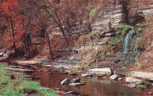 A serene stream flows beside a rocky cliff, surrounded by vibrant autumn foliage and a small waterfall.