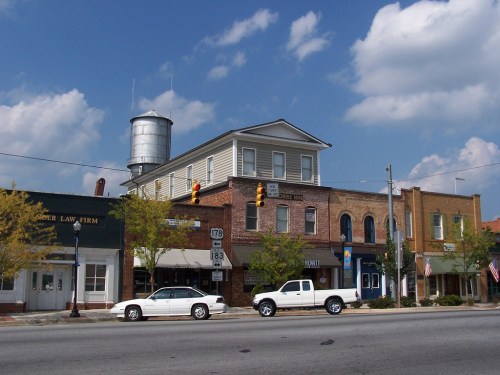 A small town street scene featuring brick buildings, a water tower, and parked cars under a partly cloudy sky.