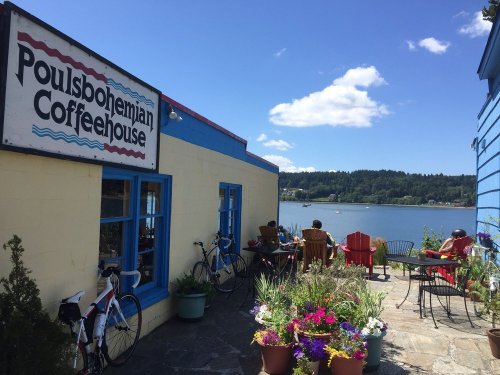 A colorful coffeehouse by the water, with outdoor seating and bicycles parked nearby, surrounded by vibrant flowers.