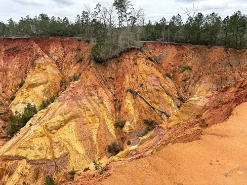 A colorful, eroded landscape with steep, layered cliffs and sparse trees against a cloudy sky.