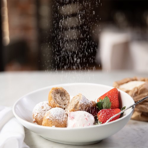 A bowl of dessert with powdered sugar being sprinkled over fried dough balls, strawberries, and mint leaves.
