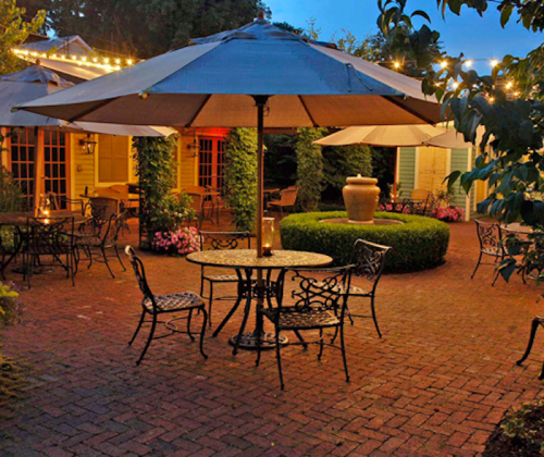 A cozy outdoor patio with tables, chairs, and umbrellas, surrounded by greenery and flowers, illuminated by soft lights.