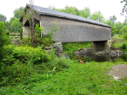 A rustic covered bridge spans a small stream, surrounded by lush greenery and trees.