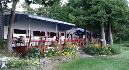 A cozy restaurant with a wooden deck, surrounded by flowers and trees, featuring outdoor seating under blue umbrellas.