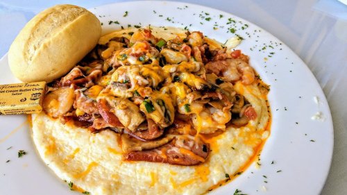 A plate of creamy grits topped with shrimp, sausage, cheese, and a roll on the side, garnished with parsley.