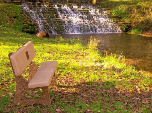A park bench sits on grass near a serene pond and a cascading waterfall in a tranquil natural setting.