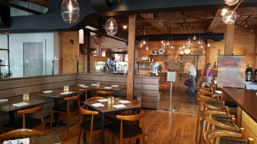 A modern restaurant interior with wooden decor, tables set for dining, and a display of kitchenware in the background.