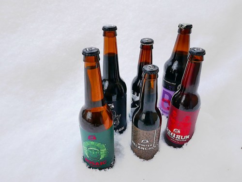 Six beer bottles with colorful labels partially buried in white snow.