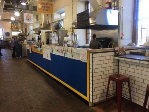 A colorful food counter in a bustling market, featuring artwork on the wall and patrons enjoying the space.