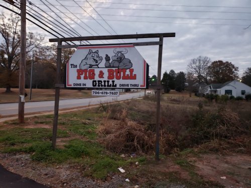 Sign for "The Pig & Bull Grill" with cartoon animals, featuring dine-in and drive-thru options, set by a road.