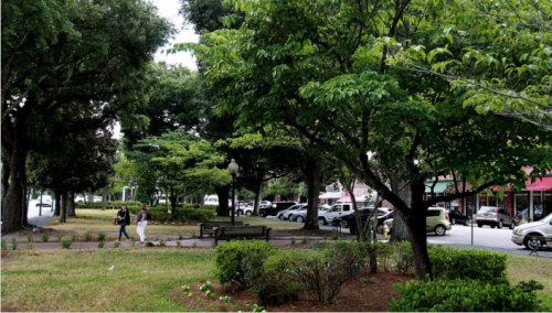 A park scene with lush green trees, a bench, and people walking along a path near parked cars and shops.