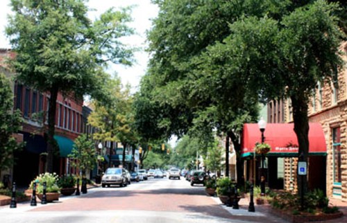 A tree-lined street with shops and parked cars, featuring brick pavement and vibrant green foliage.