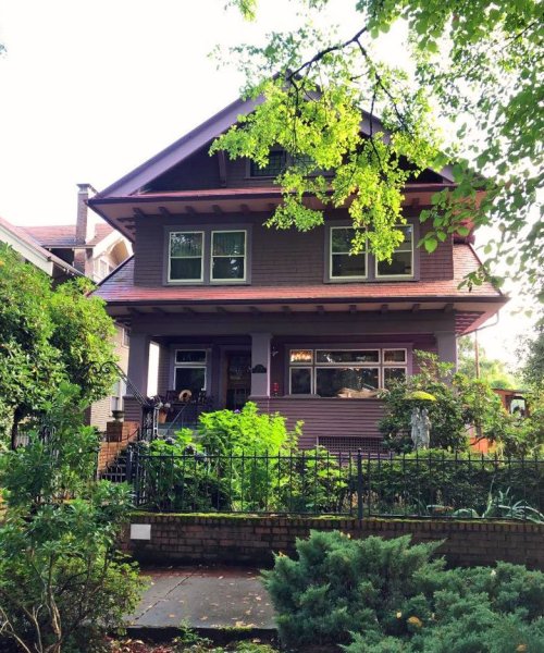 A large, two-story purple house surrounded by greenery and trees, featuring a welcoming front porch.