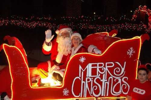 Santa Claus and a woman in a festive outfit sit in a brightly lit sleigh, waving with a "Merry Christmas" sign.