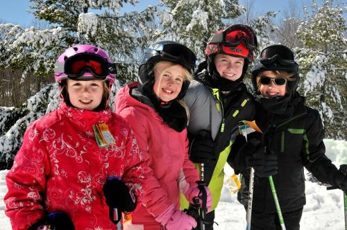 Four children in colorful ski gear smile together in a snowy landscape, surrounded by trees.