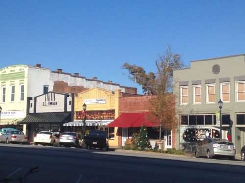 A small town street with historic buildings, parked cars, and a decorated Christmas tree under a clear blue sky.