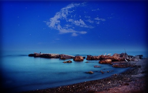 A serene beach scene at dusk, featuring calm waters, rocky shore, and a starry sky with wispy clouds.