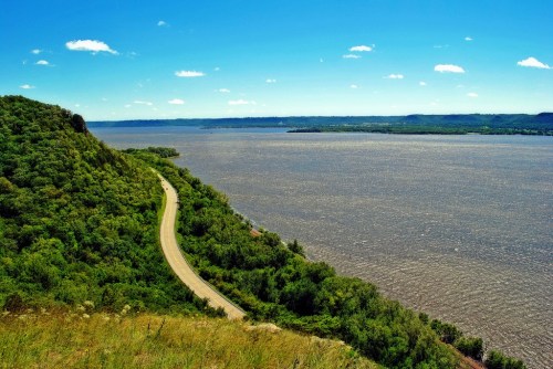 A winding road along a lush green hillside overlooking a wide river under a clear blue sky.