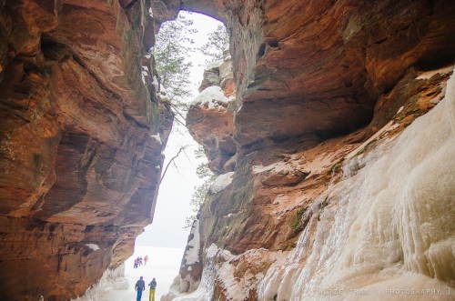 Two people walk through a snowy rock formation with an arch, surrounded by icy cliffs and trees in the background.