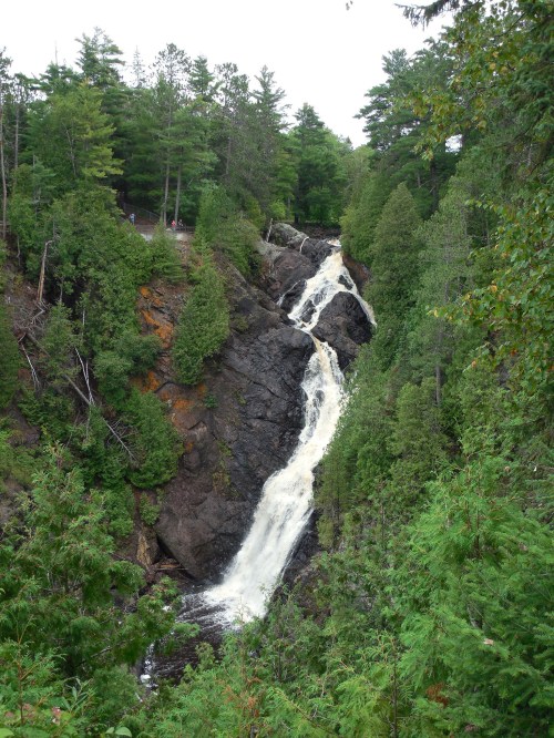 A cascading waterfall flows down rocky cliffs, surrounded by lush green trees and a serene natural landscape.