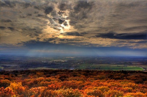 A panoramic view of a landscape with vibrant autumn foliage under a dramatic, cloudy sky.
