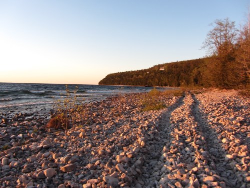 A rocky beach at sunset, with gentle waves and a forested cliff in the background.