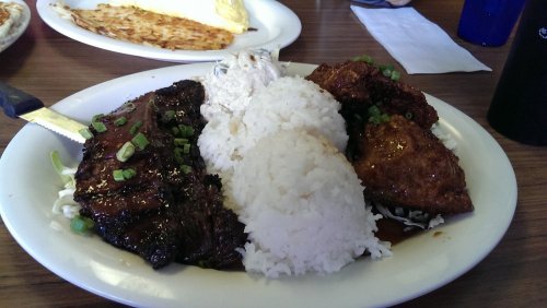 A plate of grilled meat with rice, topped with green onions, alongside a side of fried food and an omelet in the background.