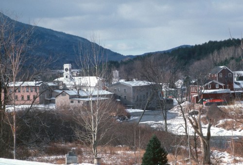 A snowy landscape featuring a small town by a river, surrounded by mountains and bare trees under a cloudy sky.