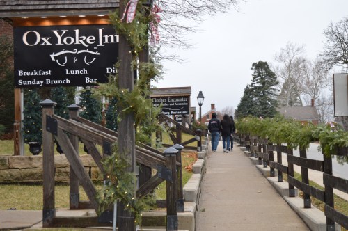 A couple walks along a sidewalk past the Ox Yoke Inn, decorated for the holidays, with trees and festive greenery.
