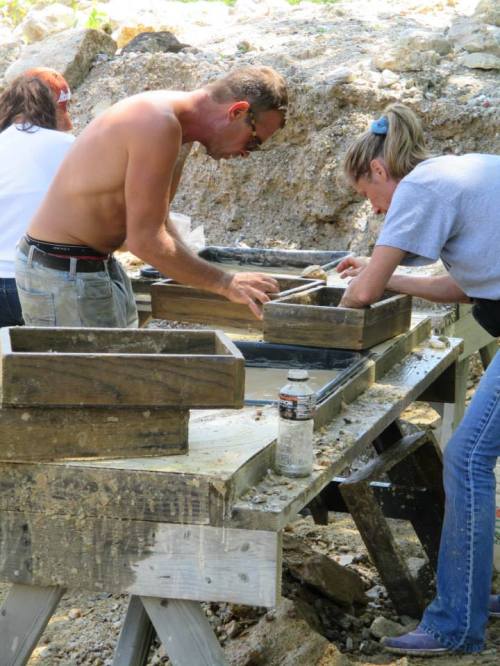 Two people sift through materials at a table outdoors, focused on their work, with a third person in the background.