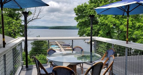 A scenic view of a lake from a deck with a glass table and chairs, surrounded by green trees and umbrellas.