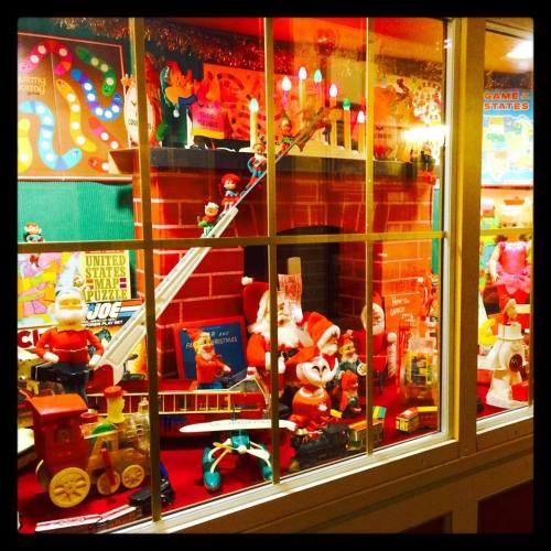 A festive display of vintage toys and decorations, featuring Santa, trains, and colorful characters behind a glass window.