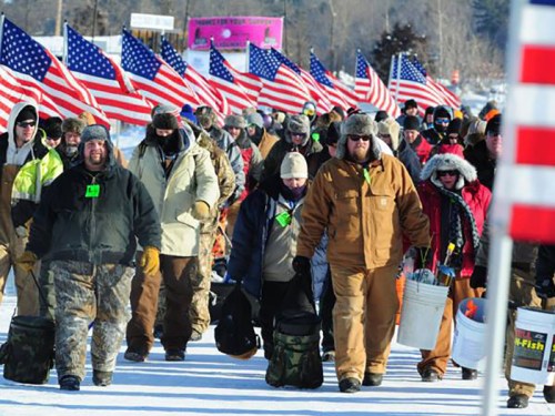 A group of people in winter clothing walking on snow, carrying gear and American flags in a festive outdoor event.