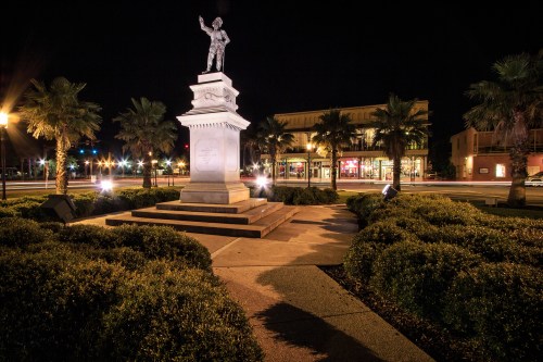 A statue on a pedestal at night, surrounded by palm trees and shrubs, with city lights in the background.
